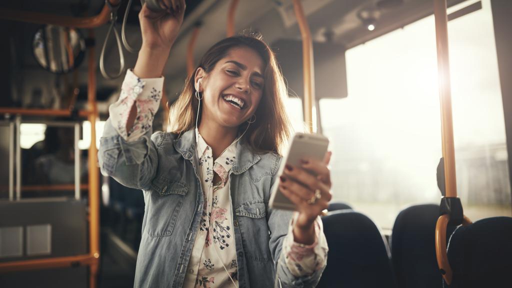 Young woman wearing earphones laughing at a text message on her cellphone while riding on a bus.