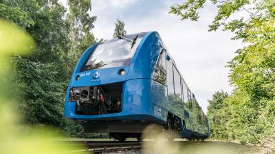 Blue passenger train powered by hydrogen running on train tracks past green foliage
