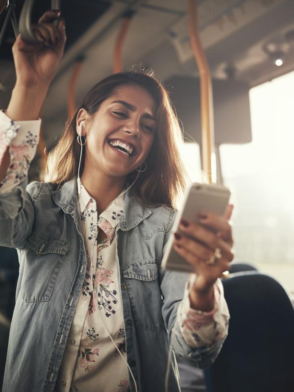 Young woman wearing earphones laughing at a text message on her cellphone while riding on a bus.