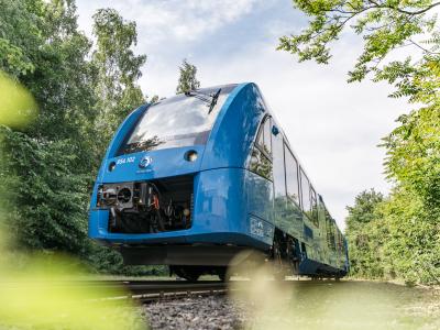 Blue passenger train powered by hydrogen running on train tracks past green foliage