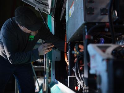 A technician wearing a gray cap holding a flashlight while looking at electric components in a bus