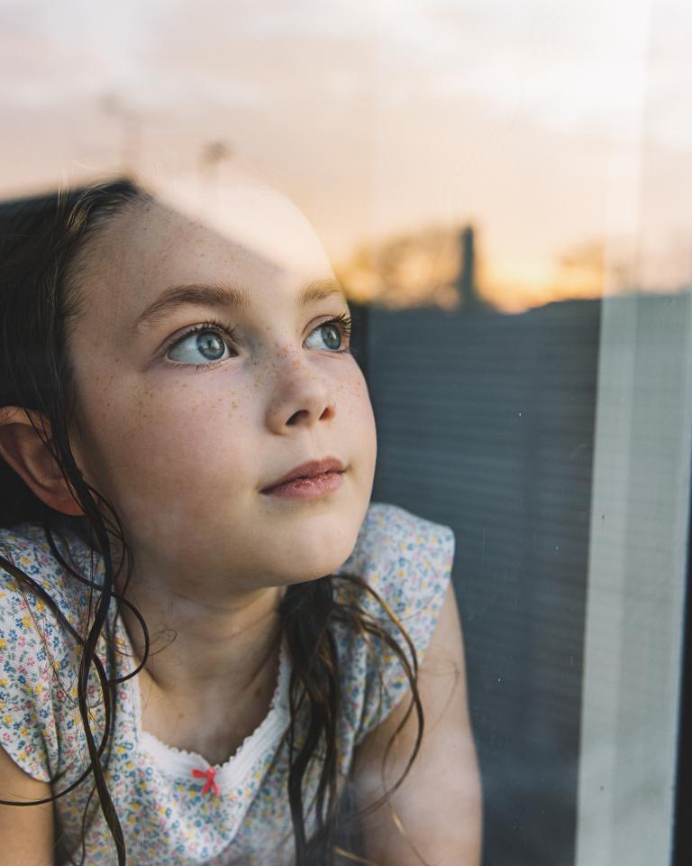 Junges Mädchen, das bei Sonnenuntergang durch das Fenster schaut