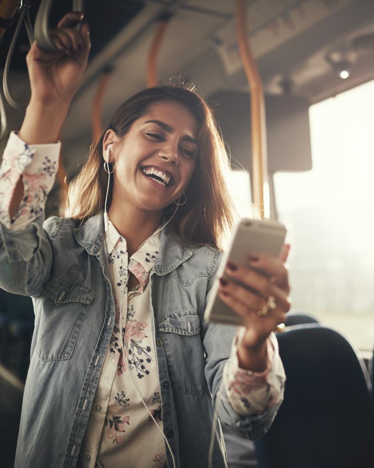 Young woman wearing earphones laughing at a text message on her cellphone while riding on a bus.