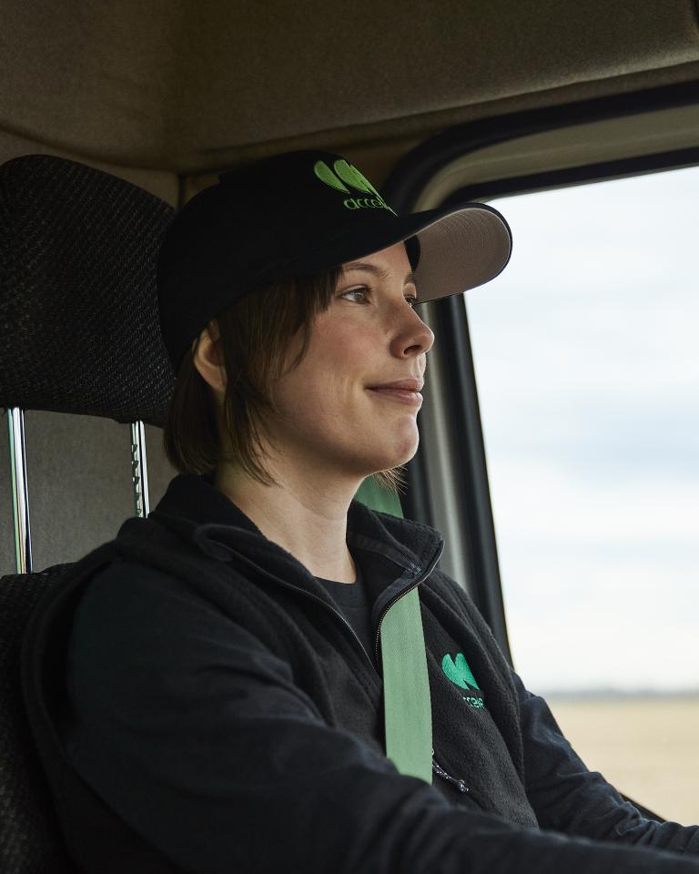 Close up image of a female truck driver with short hair, wearing an Accelera baseball hat, driving a zero-emission truck