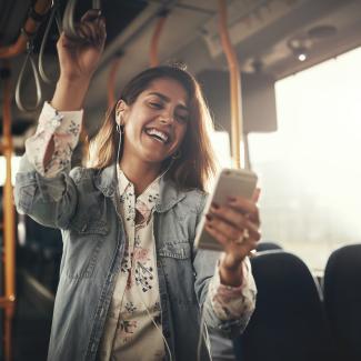 Junge Frau mit Kopfhörern lacht über eine SMS auf ihrem Handy, während sie in einem Bus fährt.